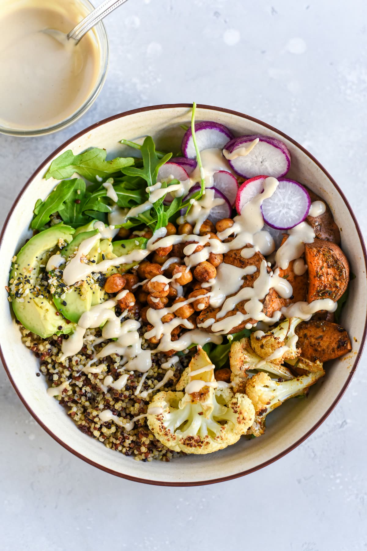 Vista de cima de um bowl com quinoa, grão-de-bico assado, abacate, rabanete, rúcula, couve-flor e batata-doce assadas, regado com molho de tahine